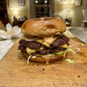 Homemade smash burger with crispy beef patties, melted cheese, pickles, shredded lettuce, onions, and creamy burger sauce on a toasted bun served on a wooden cutting board. BuyMyRecipes.com
